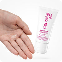 The image of a tube of Conceive Plus Fertility Lubricant 30ml on the right side of the frame while a woman left hand entering the frame from the left side on a white background. The woman hand has some lube on its palm with fingers closed.